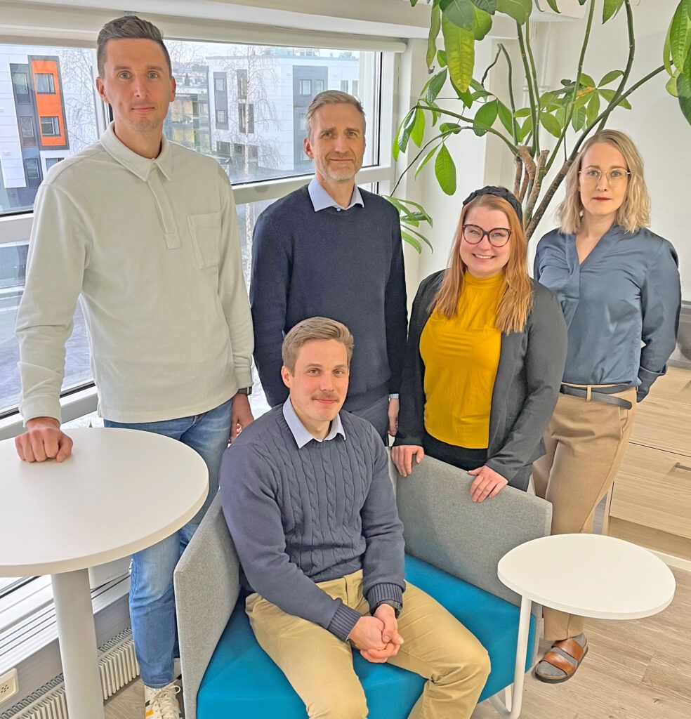 Group of five people posing together indoors; two standing, three seated.