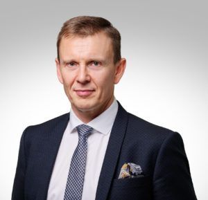 Man in suit and tie, smiling slightly, with a pocket square; studio portrait.