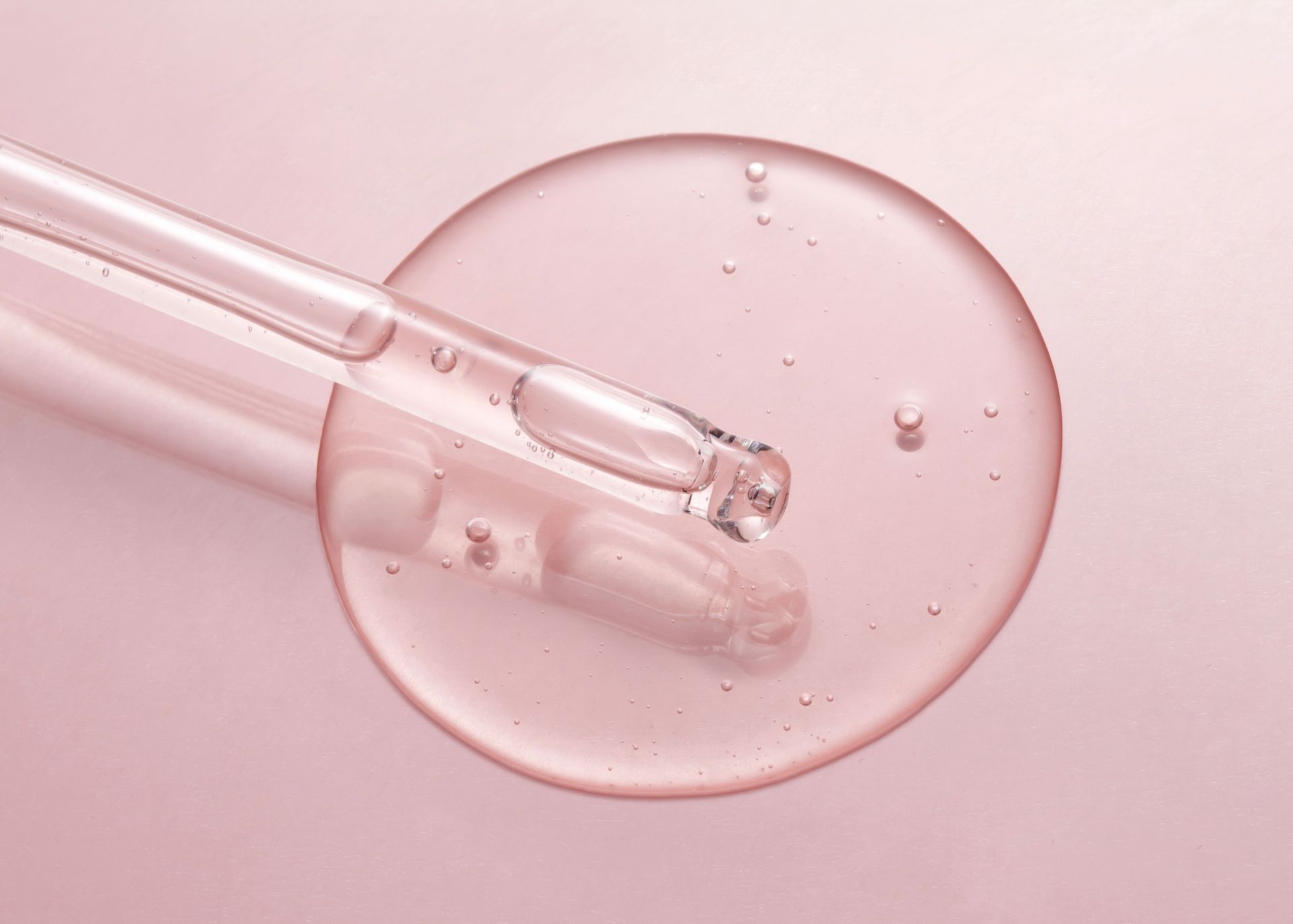 A dropper dispenses clear serum onto a pink surface, forming a droplet.