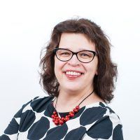 Woman with glasses smiles, wearing a black and white patterned top and red beaded necklace.