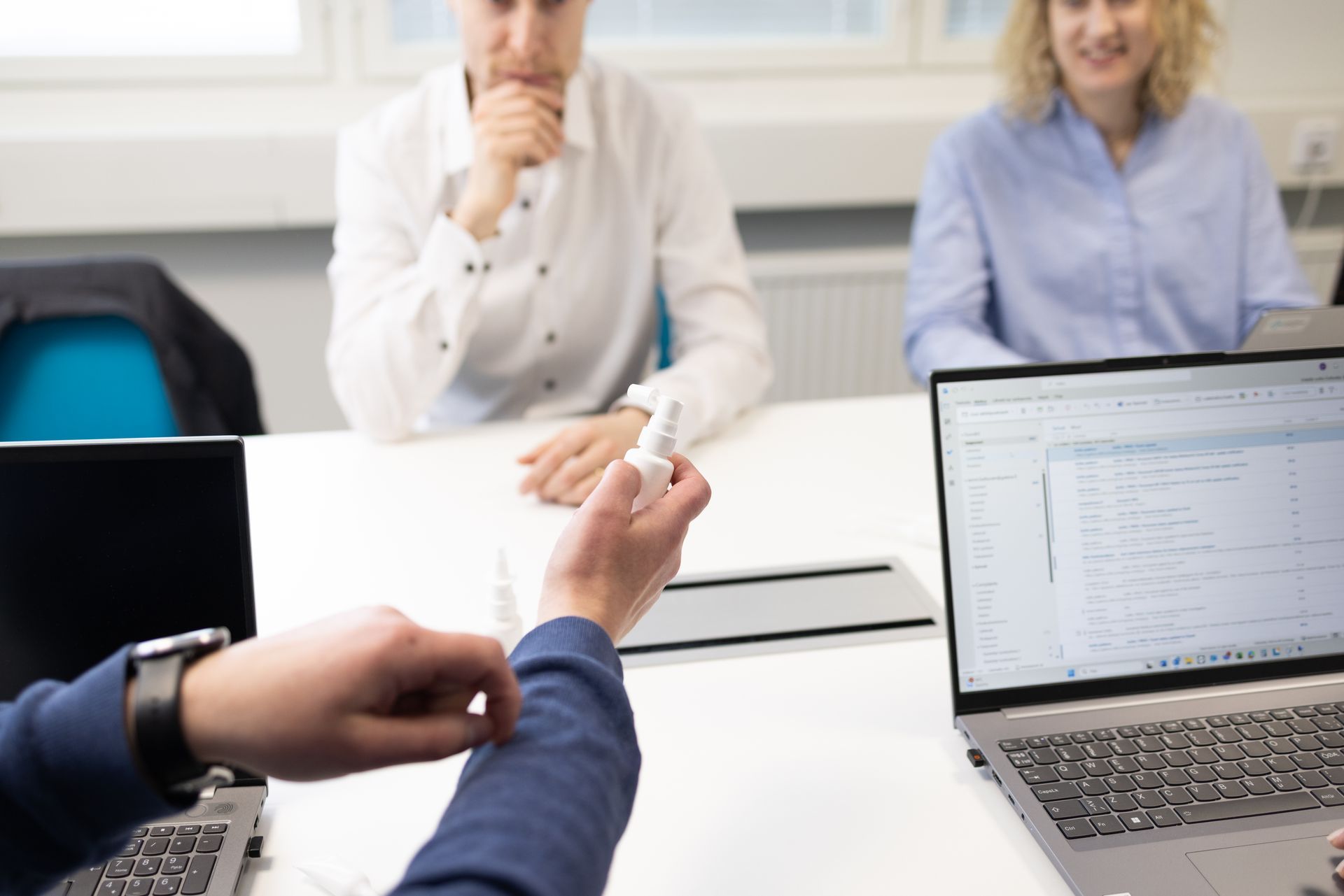 Person spraying hand sanitizer at a meeting table with two others looking on.