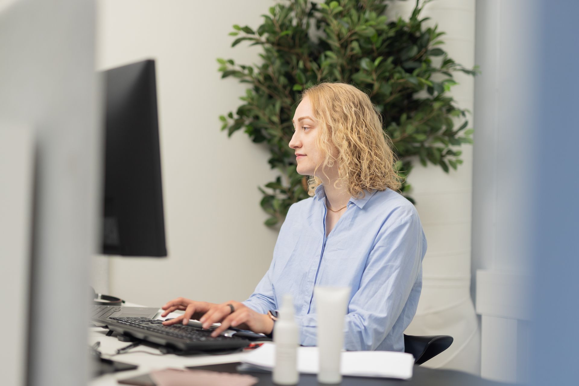 Woman with curly blonde hair typing on a keyboard, looking at a monitor, in an office.