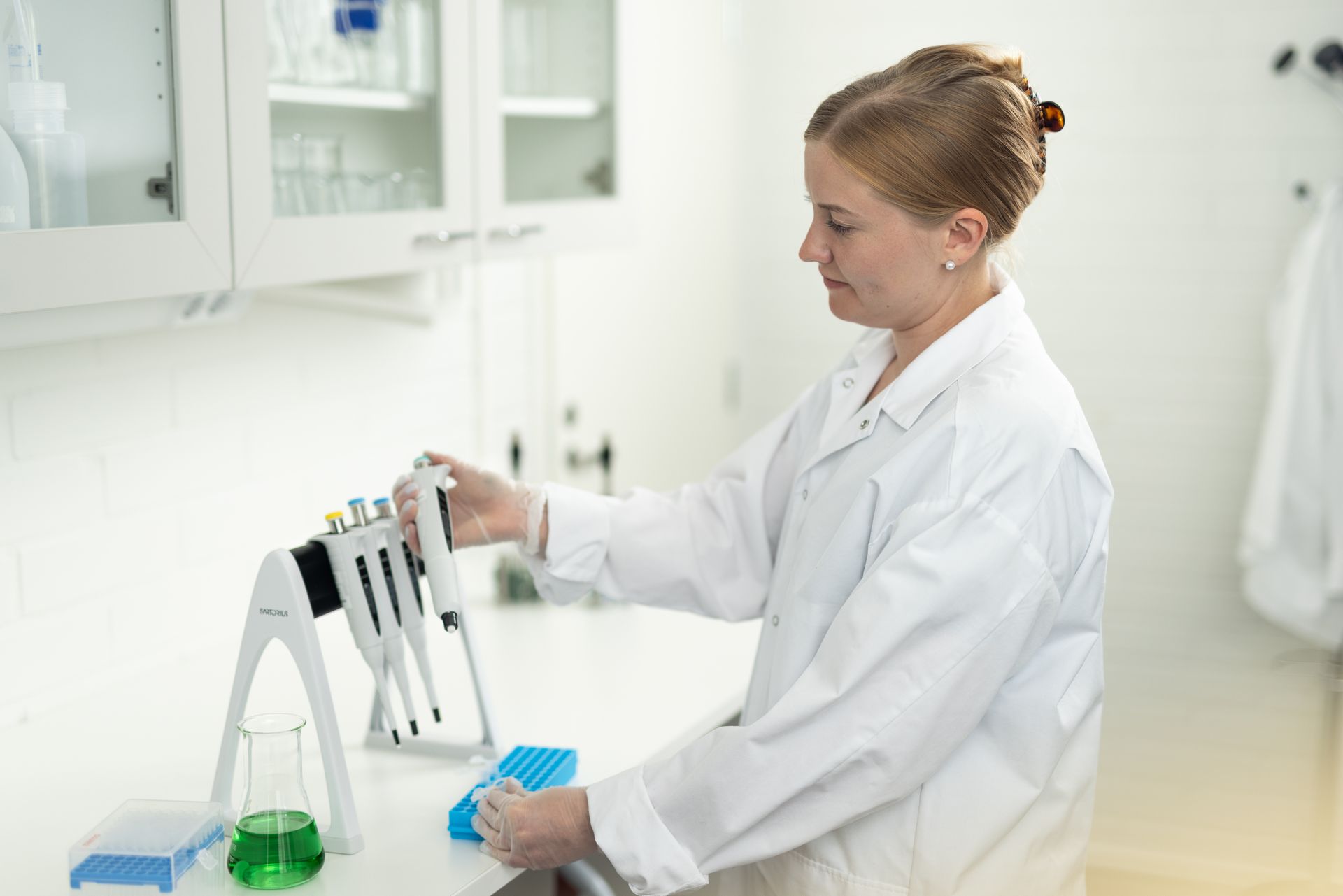 Woman in lab coat using a pipette in a laboratory setting with beakers and containers.