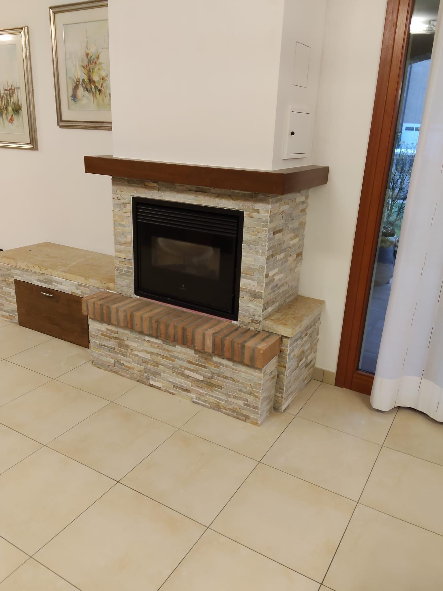 Camino del soggiorno con cornice in pietra, mensola in legno e pavimento piastrellato, accanto a una tenda e un'opera d'arte a parete.
