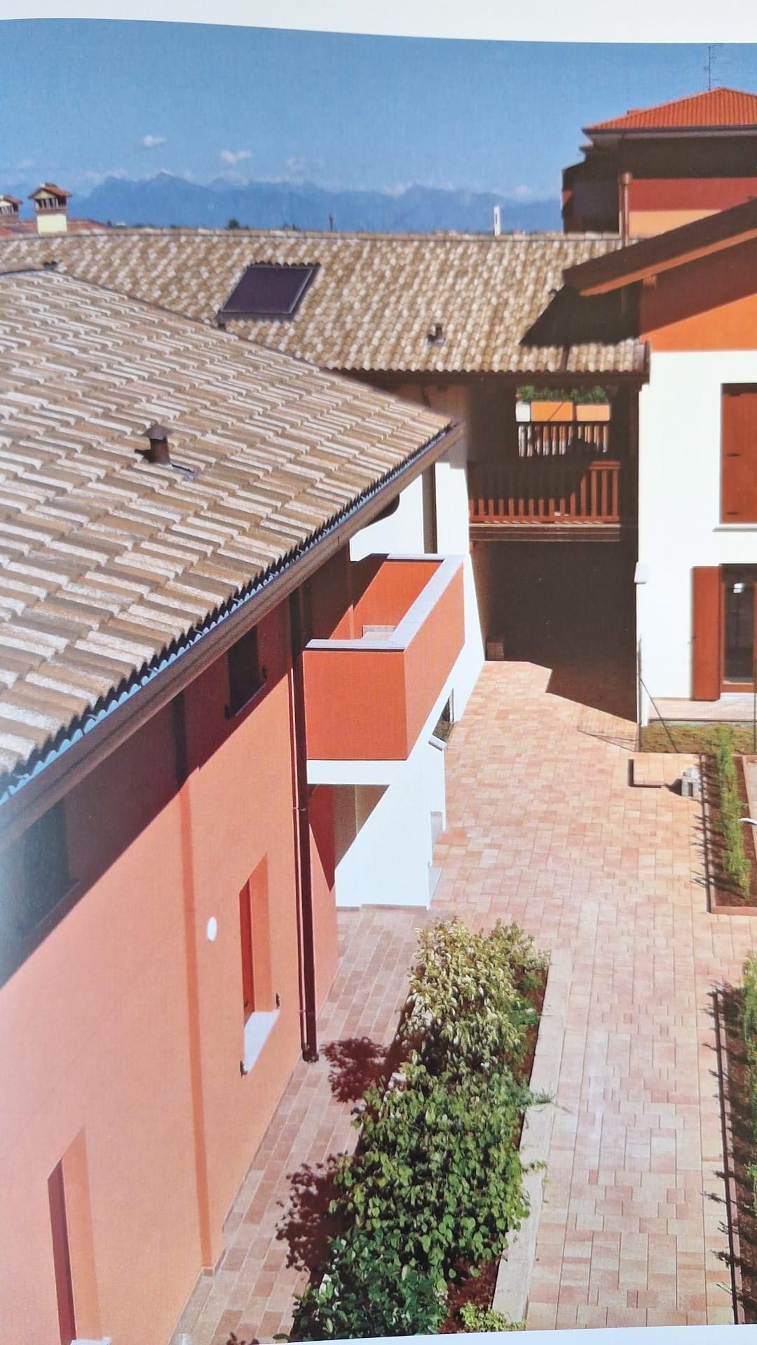 Cortile soleggiato tra edifici in terracotta con tetti di tegole e montagne in lontananza.