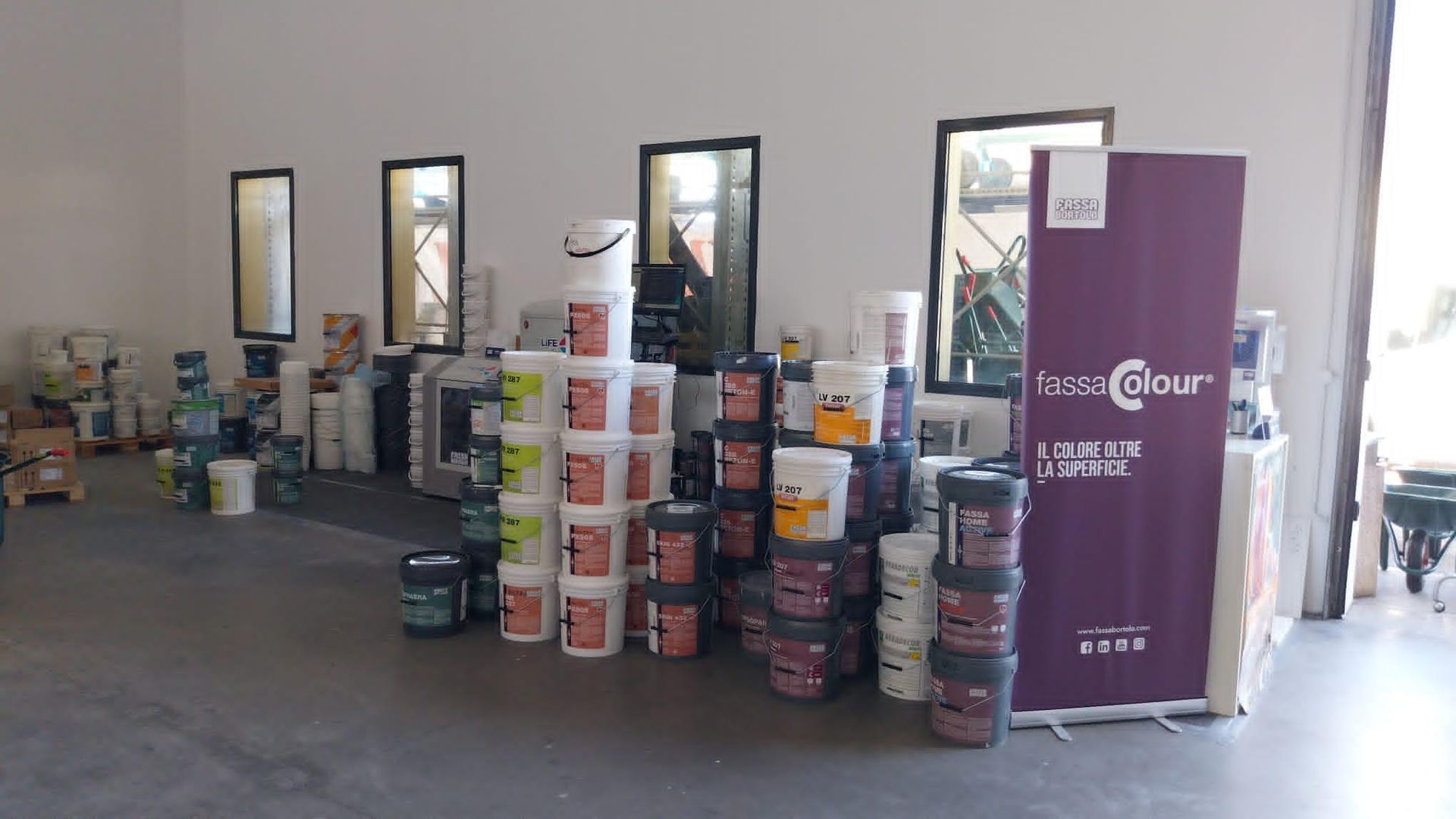 Interno del negozio con secchi di vernice impilati e materiali vicino alle finestre, tra cui un alto espositore viola