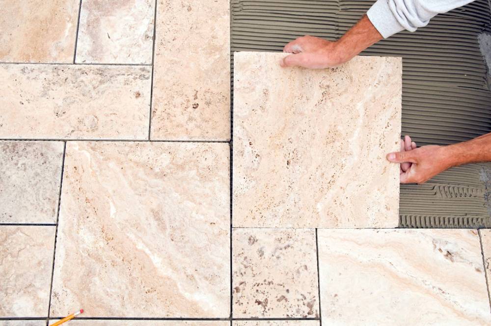 Mani che posano piastrelle di pietra beige sulla superficie di un patio esterno.