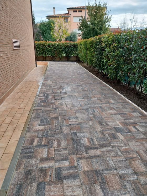 Cortile laterale pavimentato in mattoni, adiacente a un muro beige, delimitato da siepi e che conduce a una casa in lontananza.
