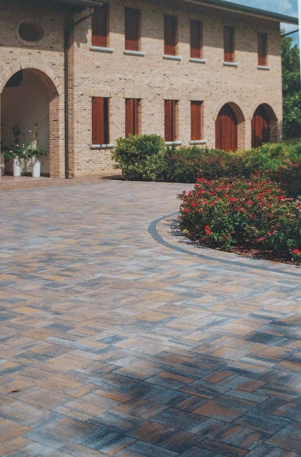 Cortile in mattoni con pavimentazione in pietra curva, facciata dell'edificio ad arco e arbusti con fiori rossi.