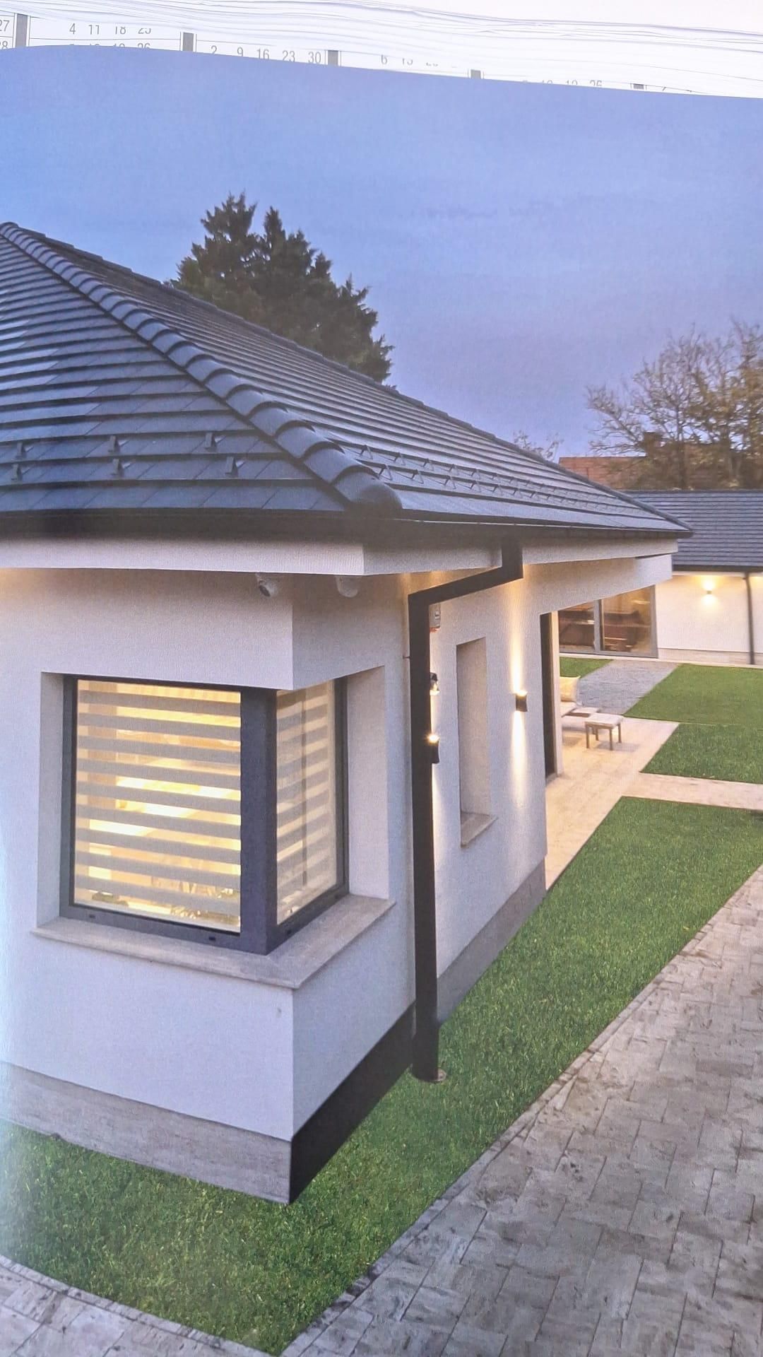 Esterno della casa con finestra illuminata, tetto di tegole e passerella accanto a un prato verde al crepuscolo