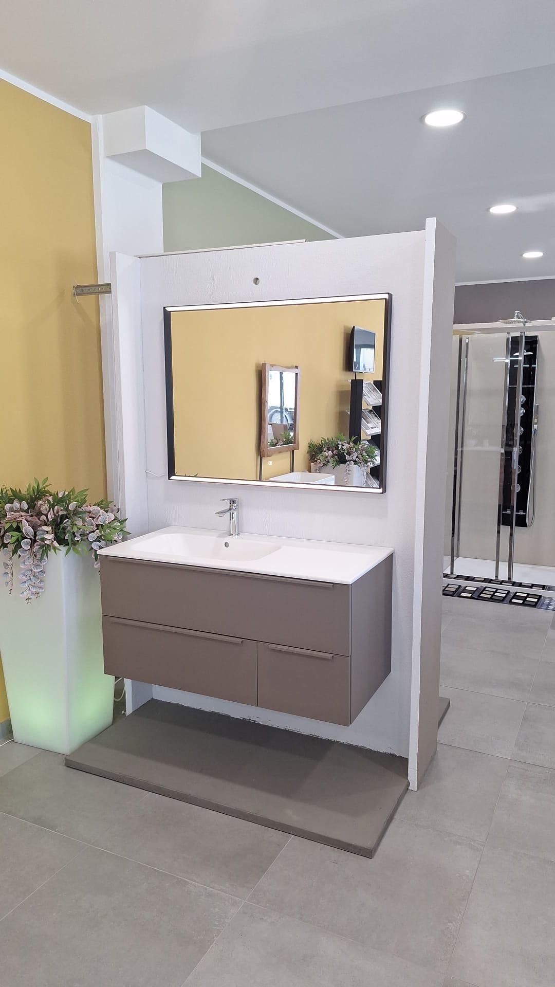 Mobile bagno moderno con specchio, pareti bianche e pavimento grigio, esposto in uno showroom.