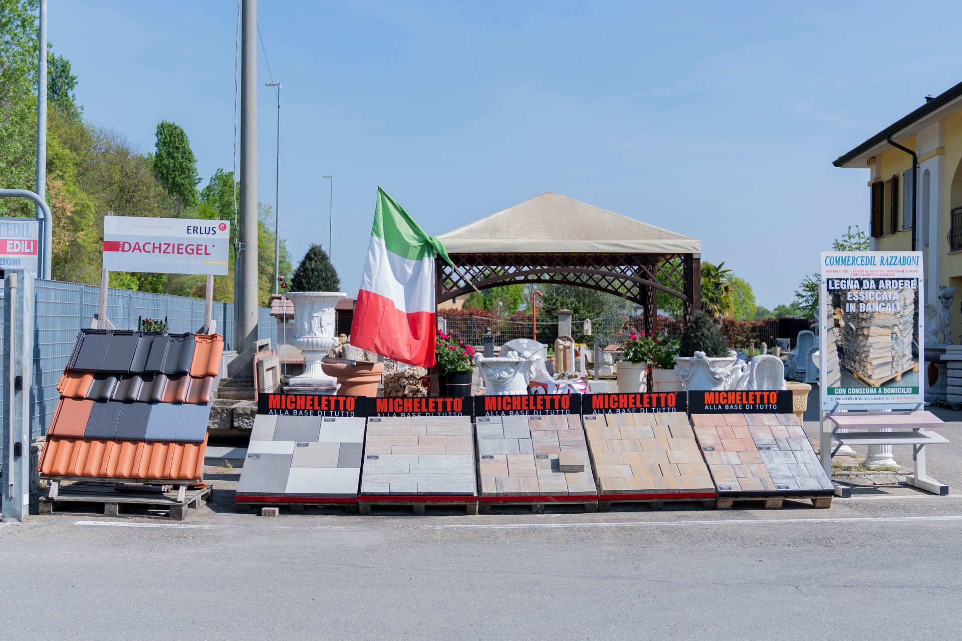 Memoriale a bordo strada con fiori, bandiere e una tettoia davanti alle barricate sotto un cielo azzurro e limpido.