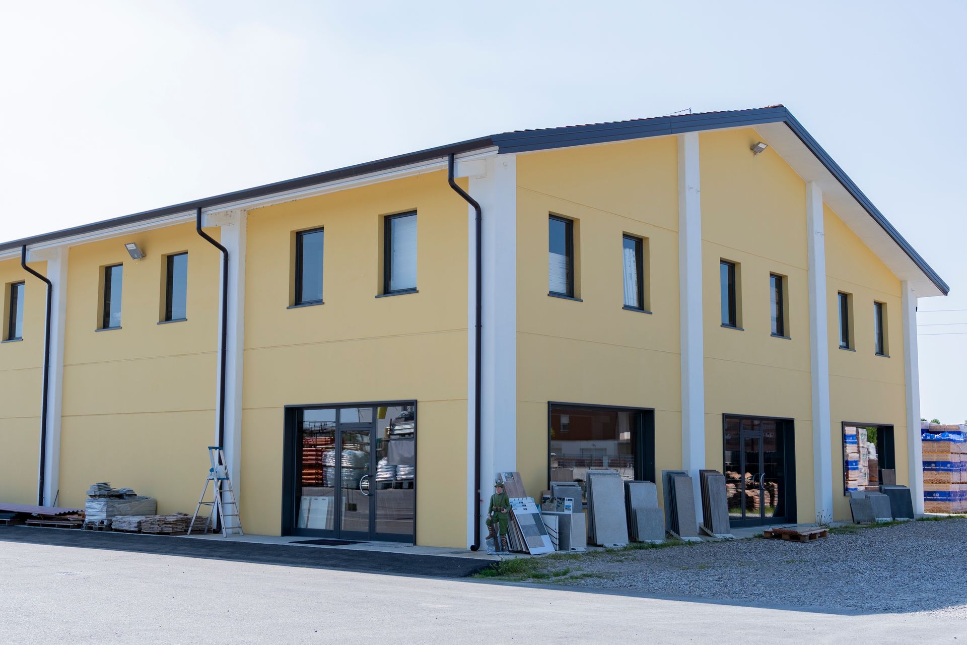 Moderno edificio commerciale giallo a due piani con ampie finestre e piazzale ghiaioso.