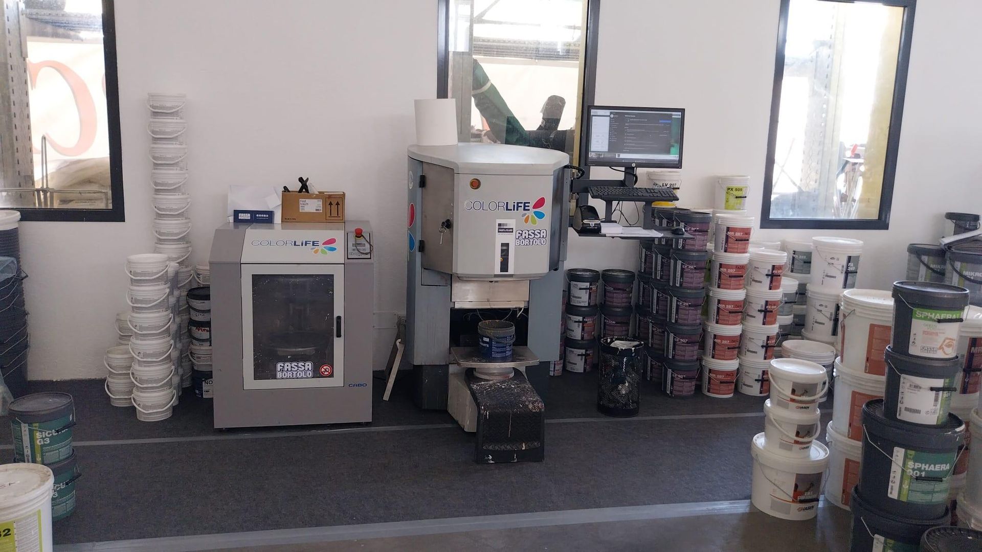 Area di lavoro industriale con macchinari, monitor per computer e contenitori di plastica impilati vicino alle finestre.