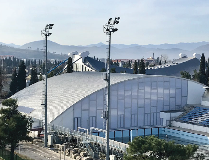Palazzetto dello sport bianco e curvo con alti riflettori, montagne sullo sfondo e tribune blu.