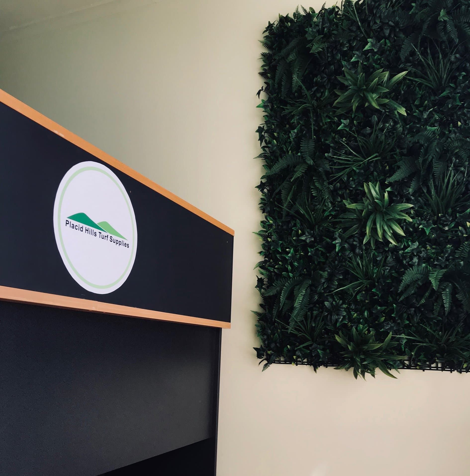Reception desk with logo, next to a green wall of faux plants. — Placid Hills Turf in Helidon, QLD