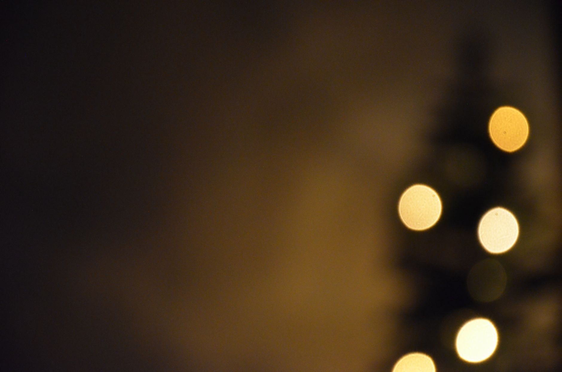 A dark, softly blurred scene of a lit tree or garland with warm glowing bokeh light circles on one side, the rest of the frame in deep shadow.