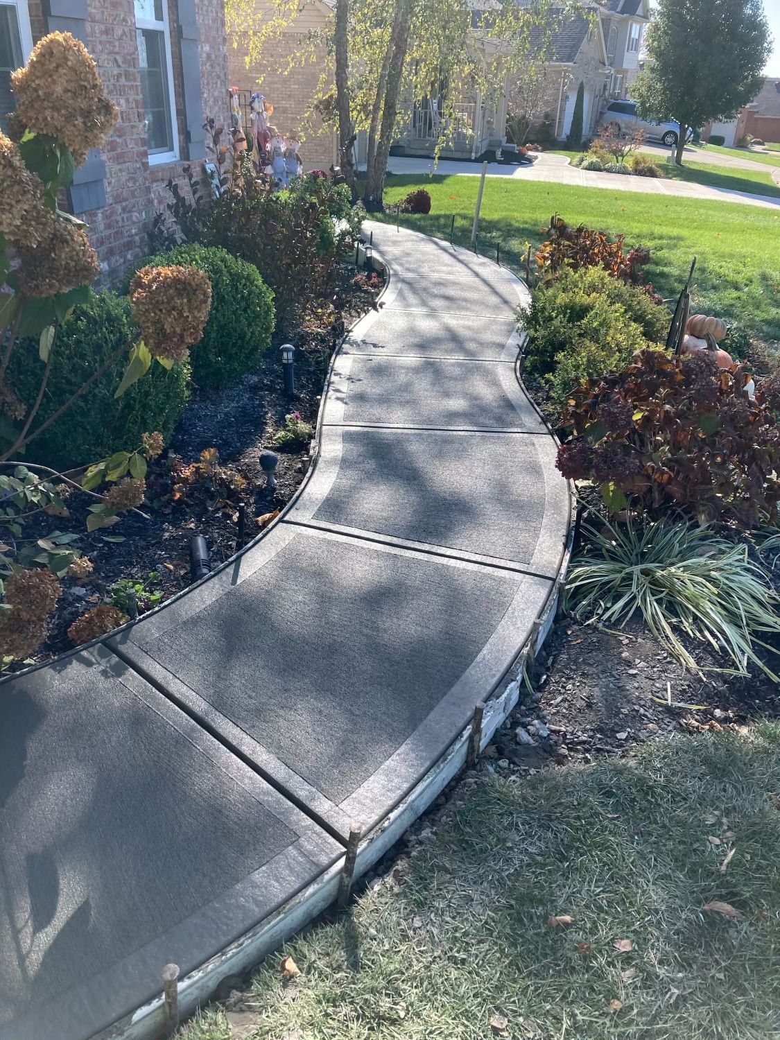 A curved concrete walkway leading to a house surrounded by flowers and bushes.