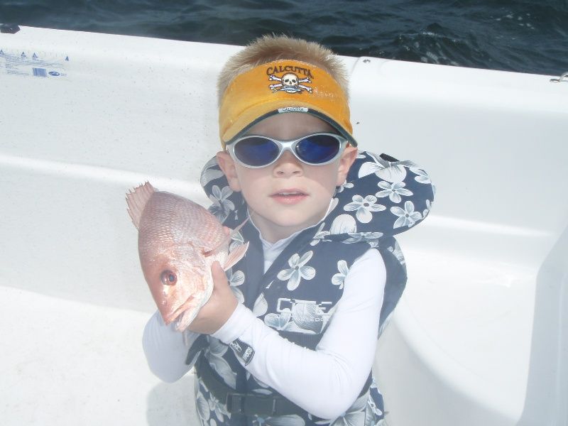 A young boy wearing a life vest and sunglasses holds a fish
