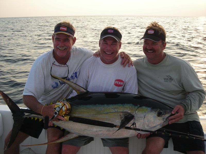 Three men on a boat holding a large fish with one wearing a shirt that says penn