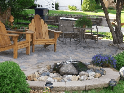 Wooden chairs on a stone patio beside a small rock fountain and blue flowers in a landscaped yard