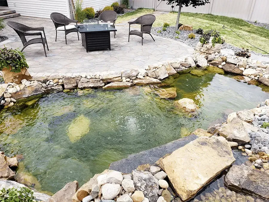 Backyard pond with rocks, patio chairs, and a fire pit on a stone deck.