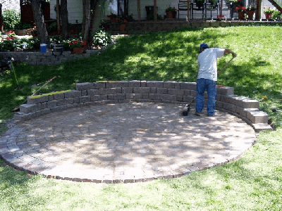 Worker building a curved stone retaining wall around a circular patio in a grassy yard.