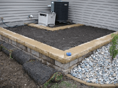 Outdoor HVAC unit on gravel beside a house, next to a raised stone-edged bed and drainage rock.