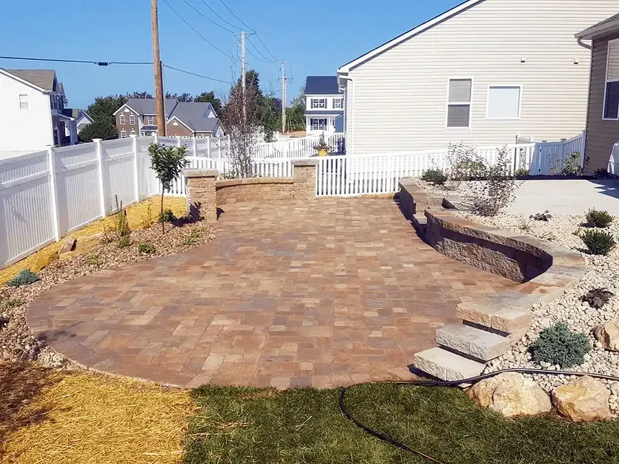 Brick patio path in a fenced backyard beside a white house, with low stone borders and mulch beds
