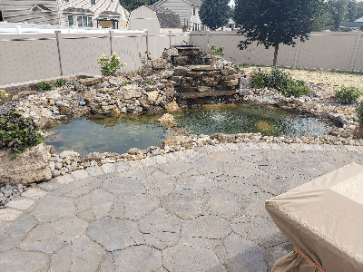 Backyard pond with rocks, waterfall, and patio seating under a fence and trees