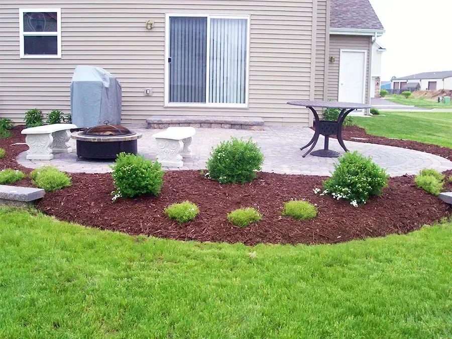 Backyard patio with potted plants, small shrubs, and a mulched flower bed beside a house