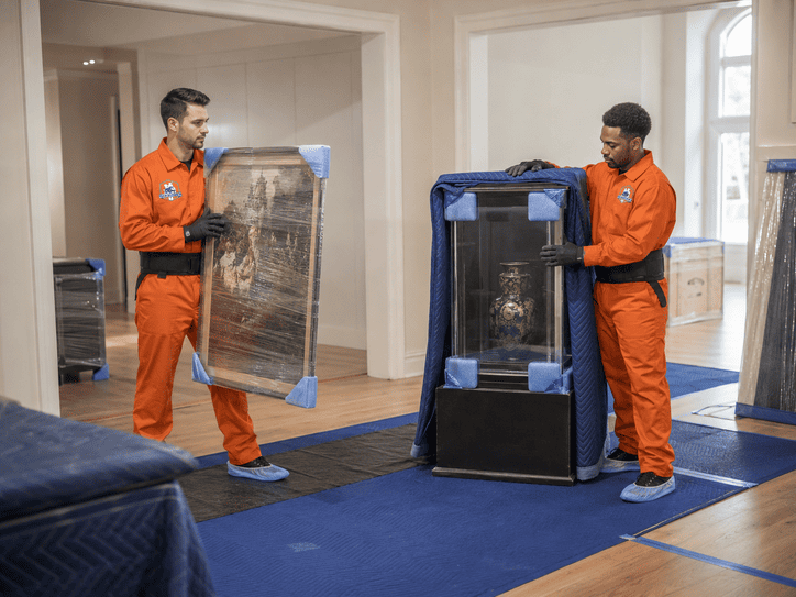 Two movers in orange uniforms carrying a wrapped appliance through a living room with blue floor runners