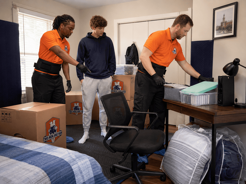 Three people moving boxes and furniture in a bedroom with a desk and chair