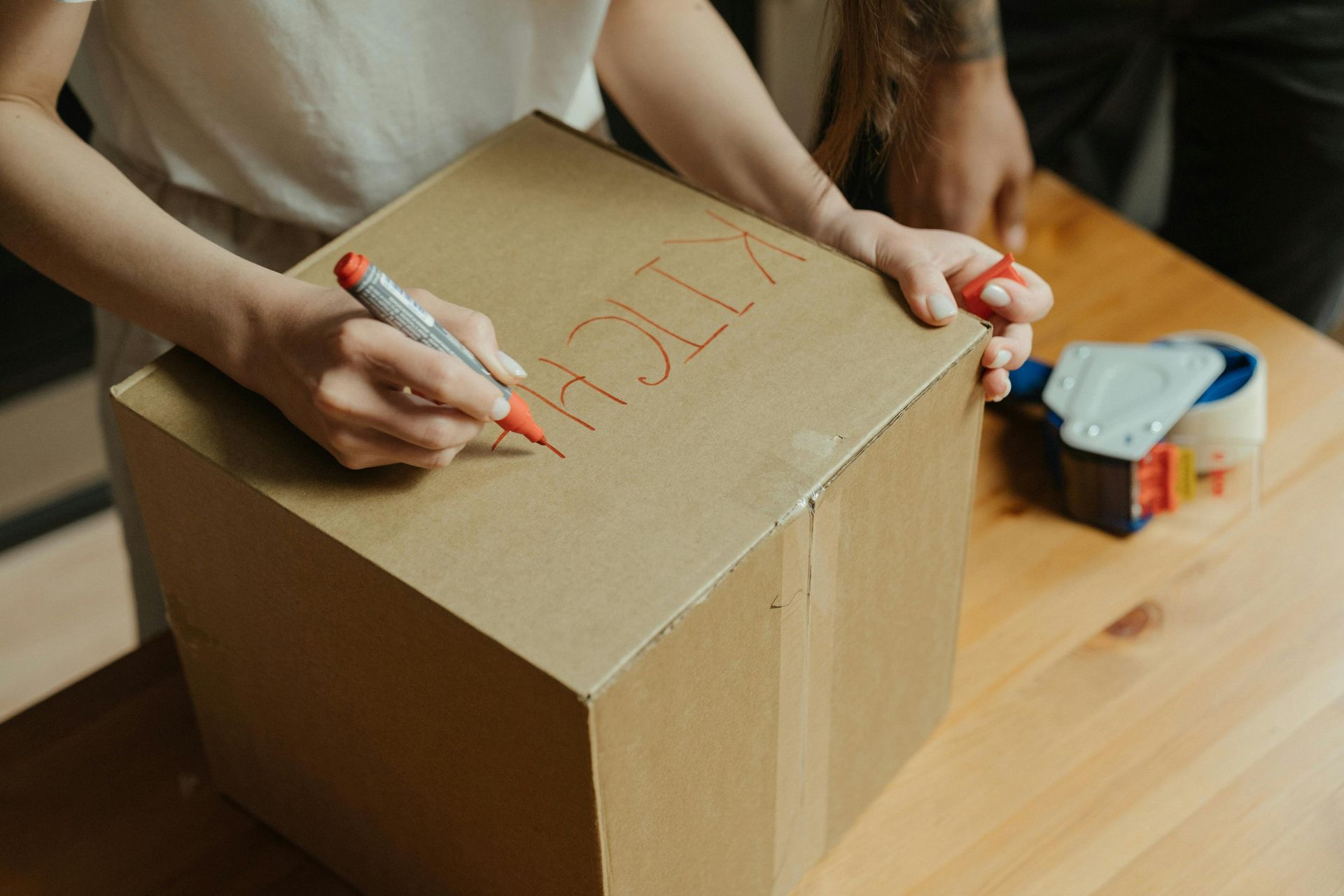 Hands writing on a cardboard box with a red marker beside packing tape and supplies on a wooden table