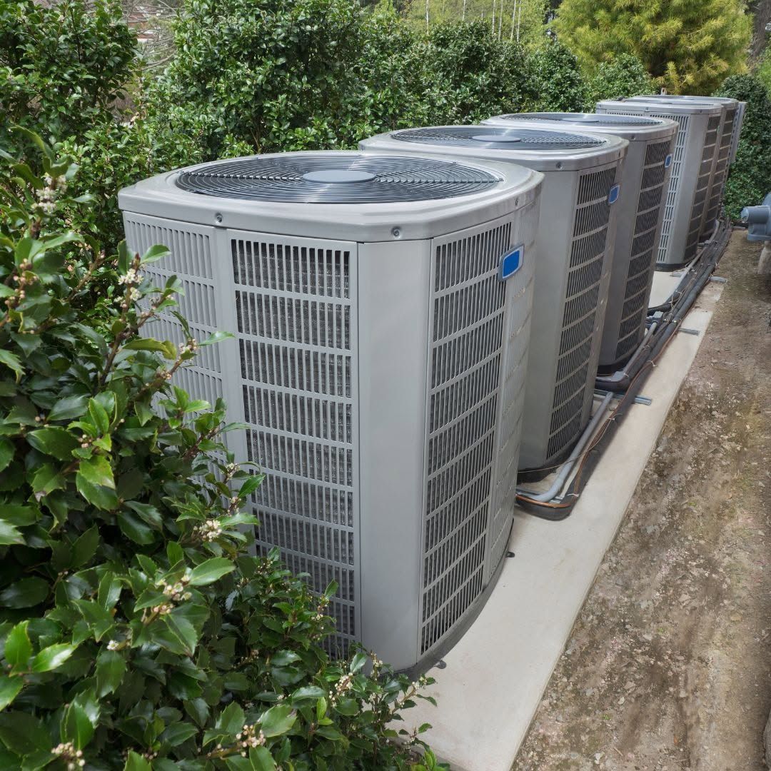 Row of gray outdoor air conditioning units along a concrete path beside dense green bushes