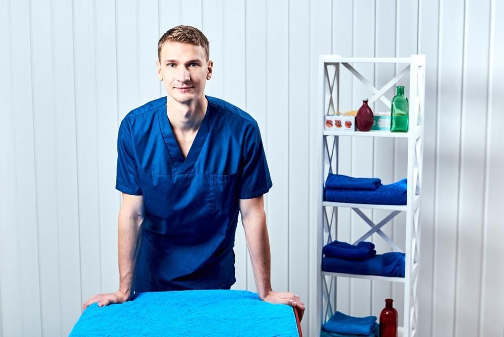 A Man In A Blue Scrub Is Standing Next To A Blue Table — Port Macquarie Physio & Pilates In Port Macquarie, NSW