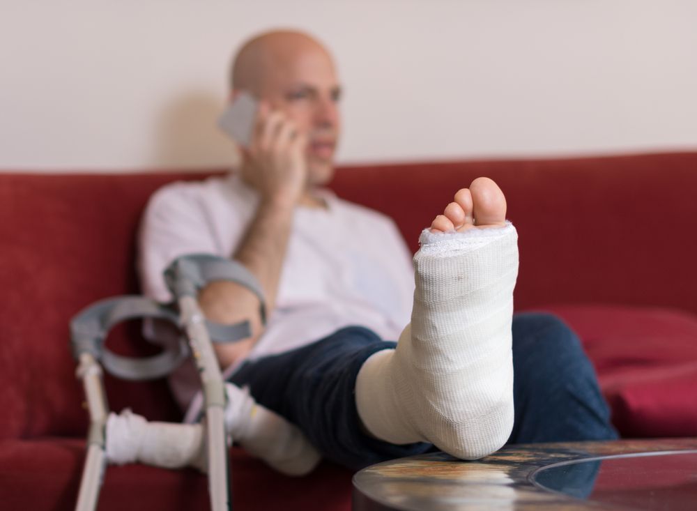 A Man With A Cast On His Leg Is Sitting On A Couch Talking On A Cell Phone — Port Macquarie Physio & Pilates In Port Macquarie, NSW