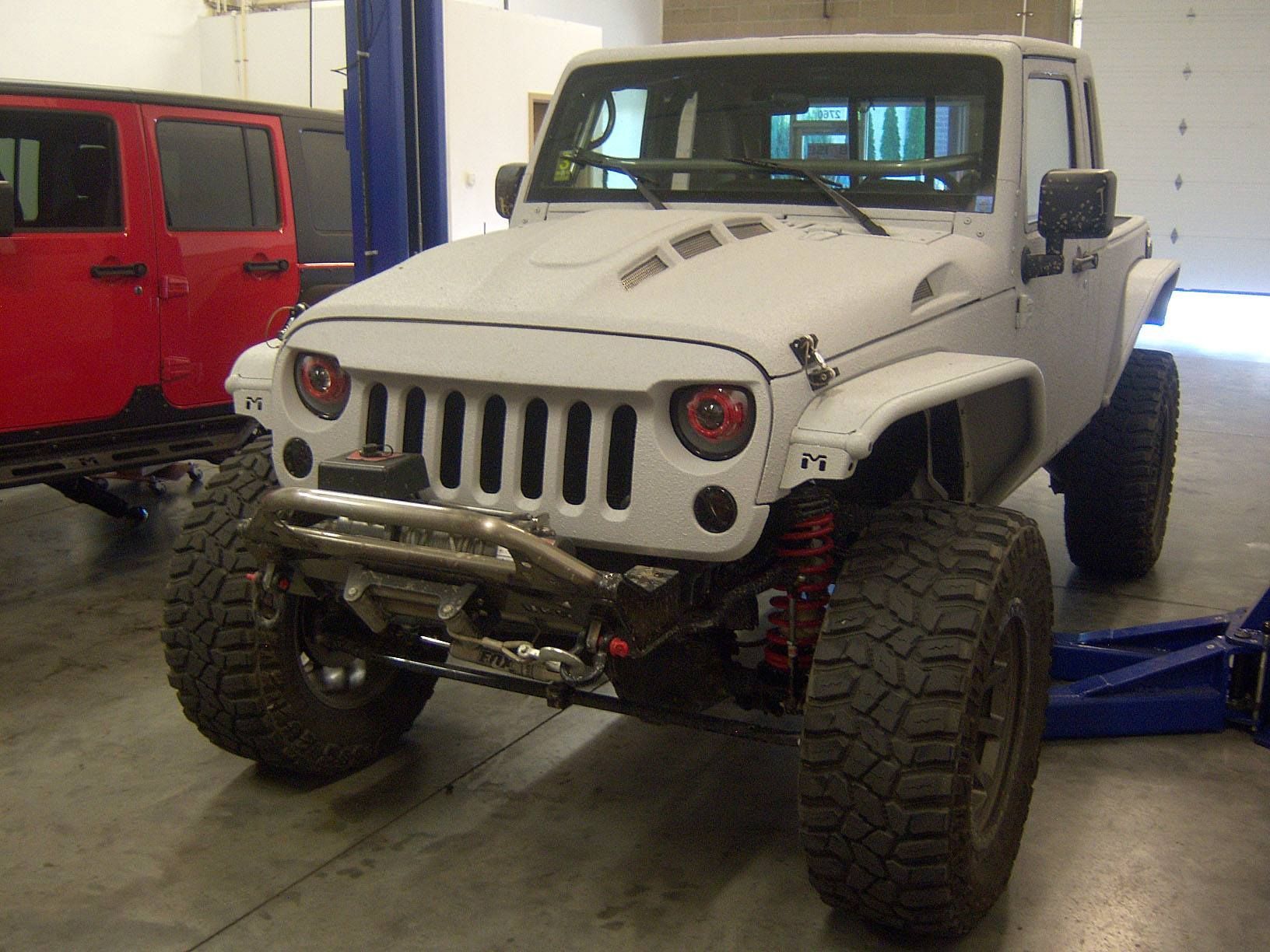 A white modified Jeep Wrangler with large off-road tires and a custom hood, parked inside a garage next to a red Jeep.
