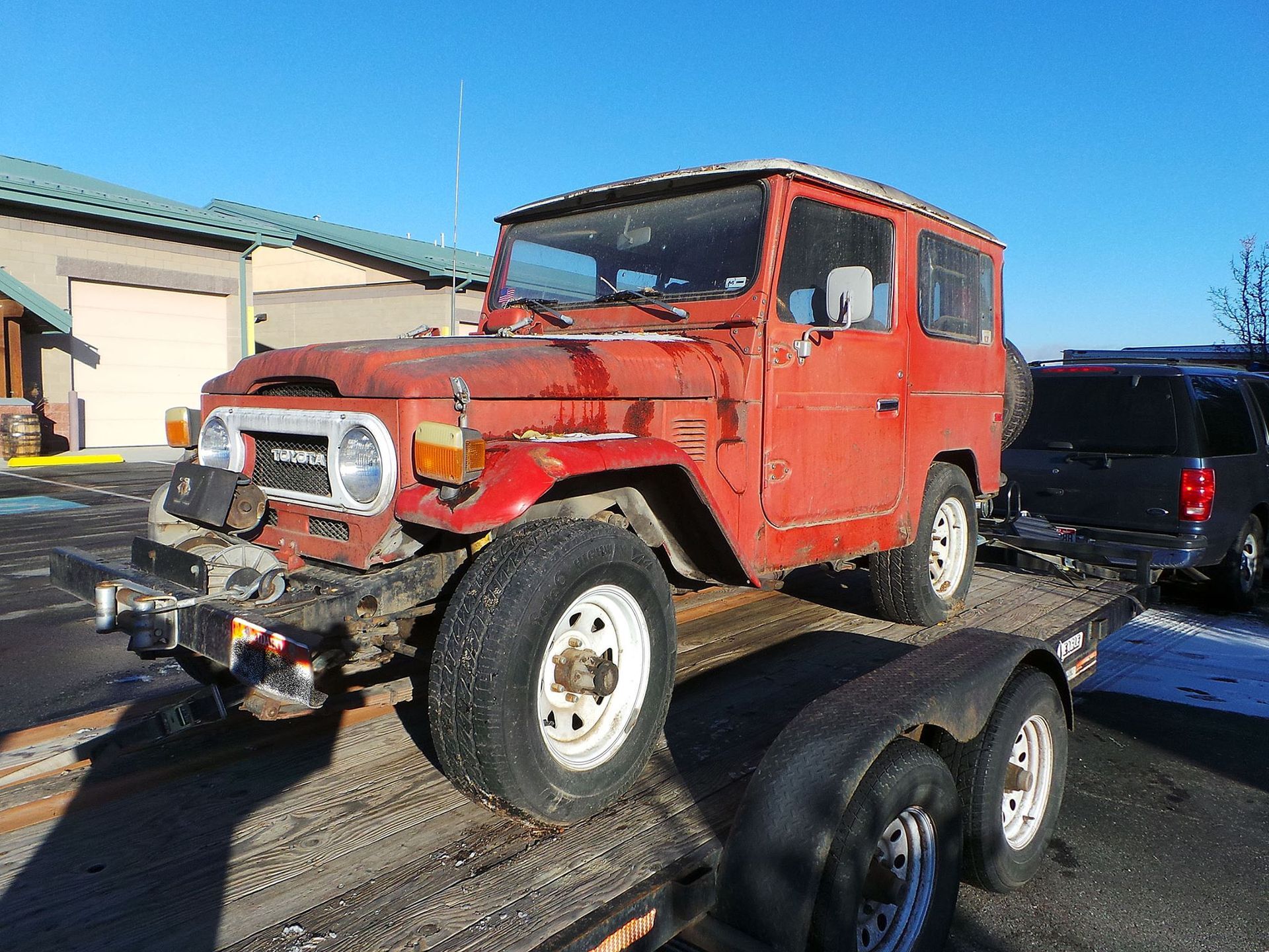 A red, weathered Toyota Land Cruiser sitting on a flatbed trailer in a parking lot on a sunny day.