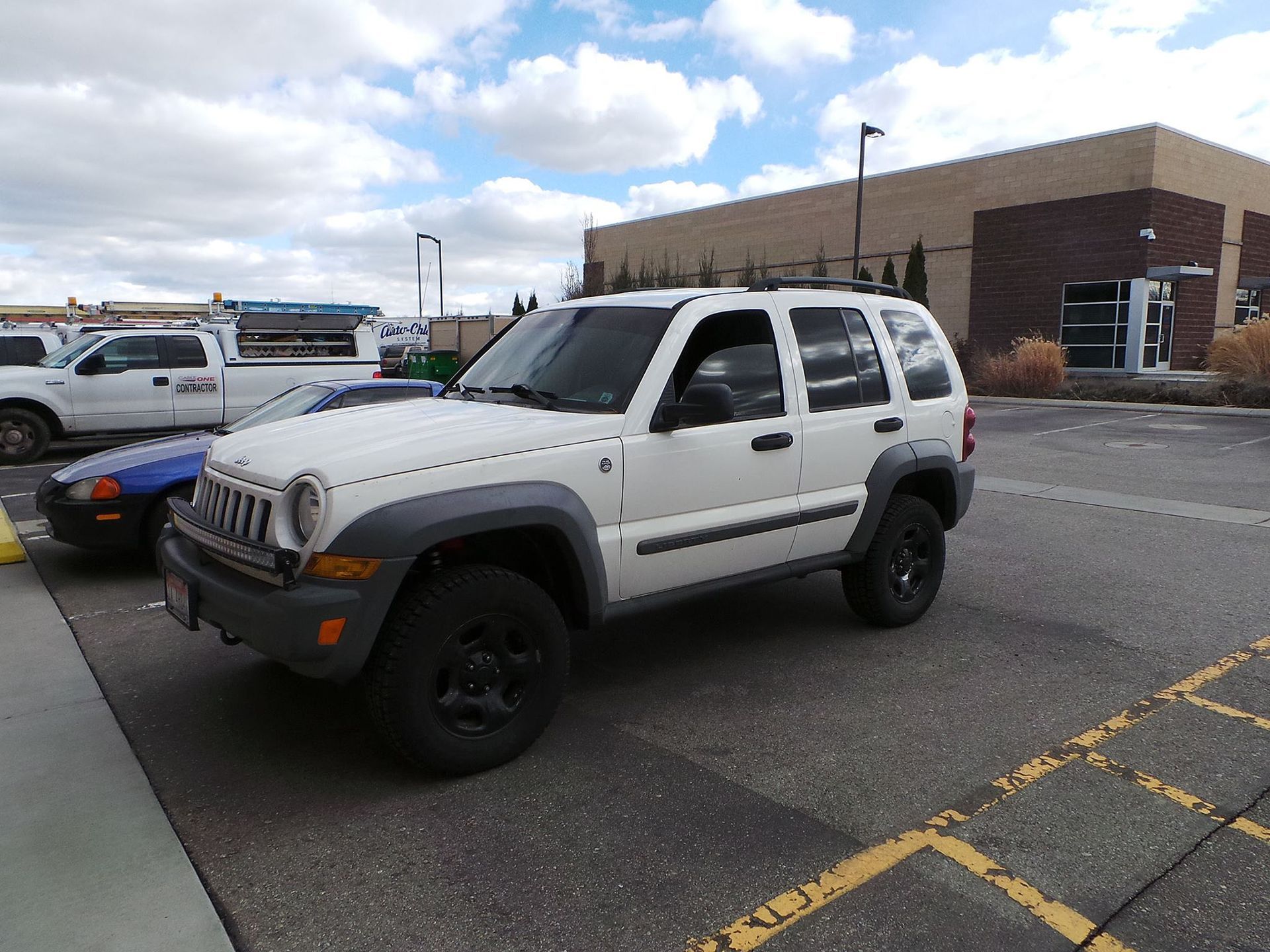 A white Jeep Liberty SUV parked in a paved parking lot on a sunny day with a building in the background.
