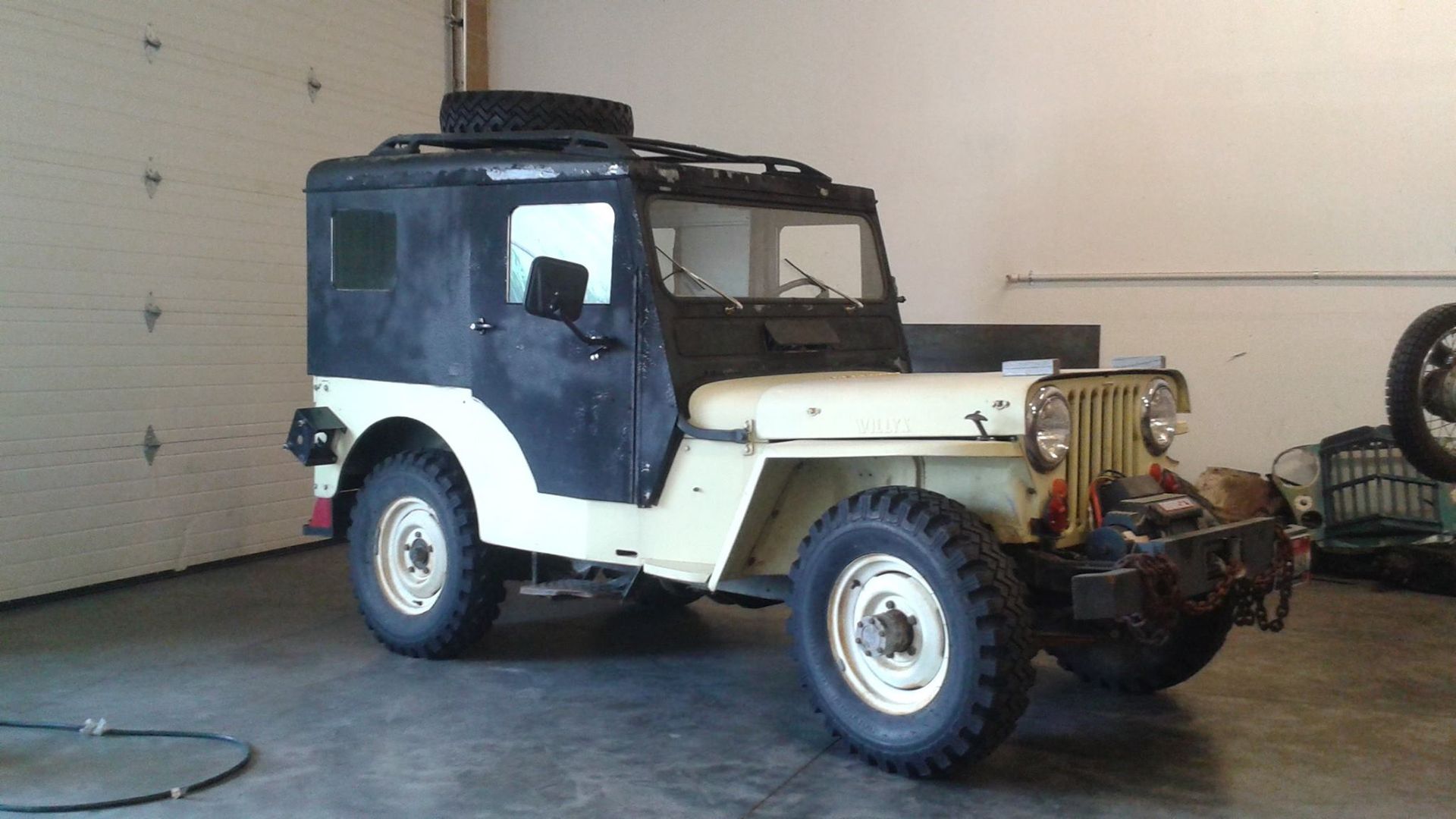 A vintage, cream-colored Jeep with a black hardtop parked inside a garage.