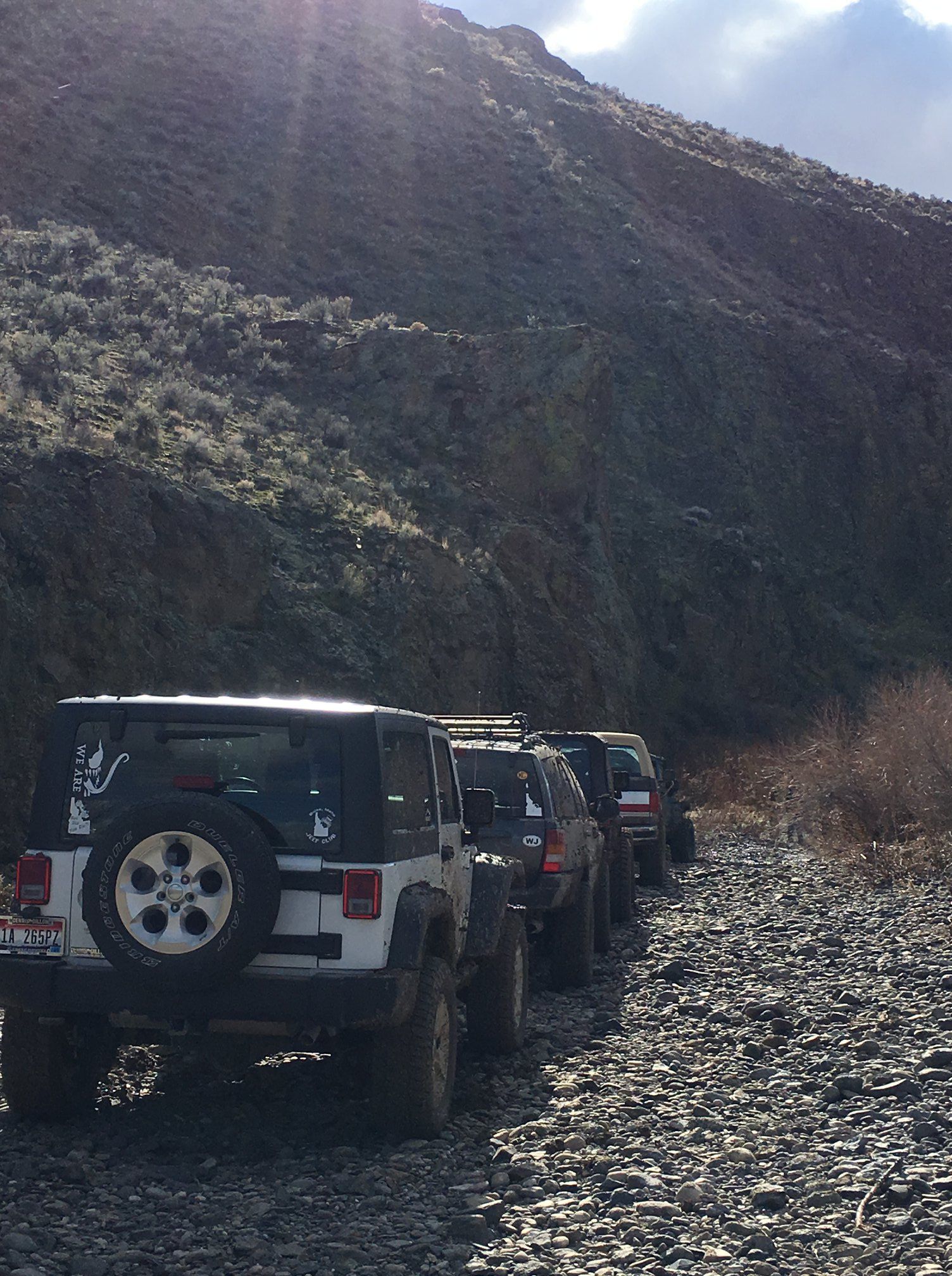 A line of off-road vehicles parked on a rocky path in front of a steep, sunlit canyon wall.