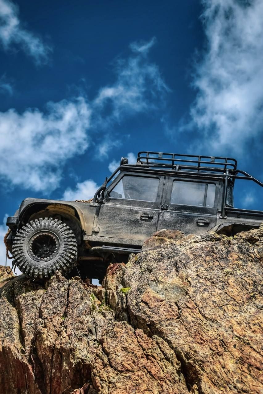 A black off-road RC truck climbs a rugged, textured rock formation against a blue, cloudy sky.