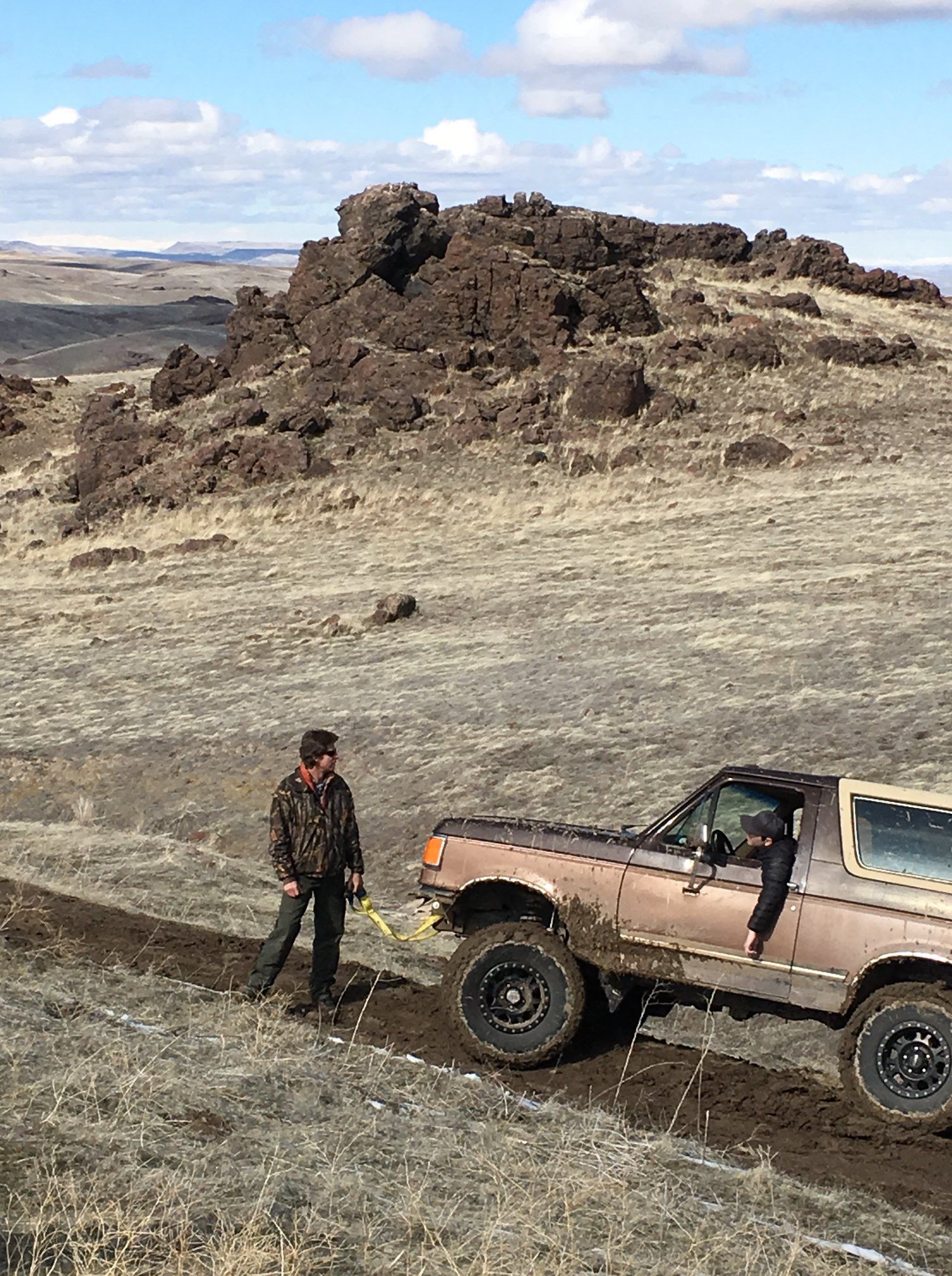 A person in camouflage clothing attaches a yellow tow strap to the front of a tan SUV stuck in a muddy, rocky landscape.