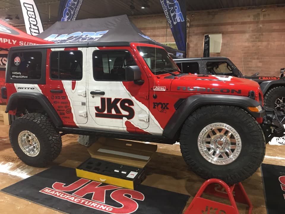 Red and white JKS Manufacturing Jeep Wrangler parked on display indoors with a prominent JKS floor mat and red jack stand.