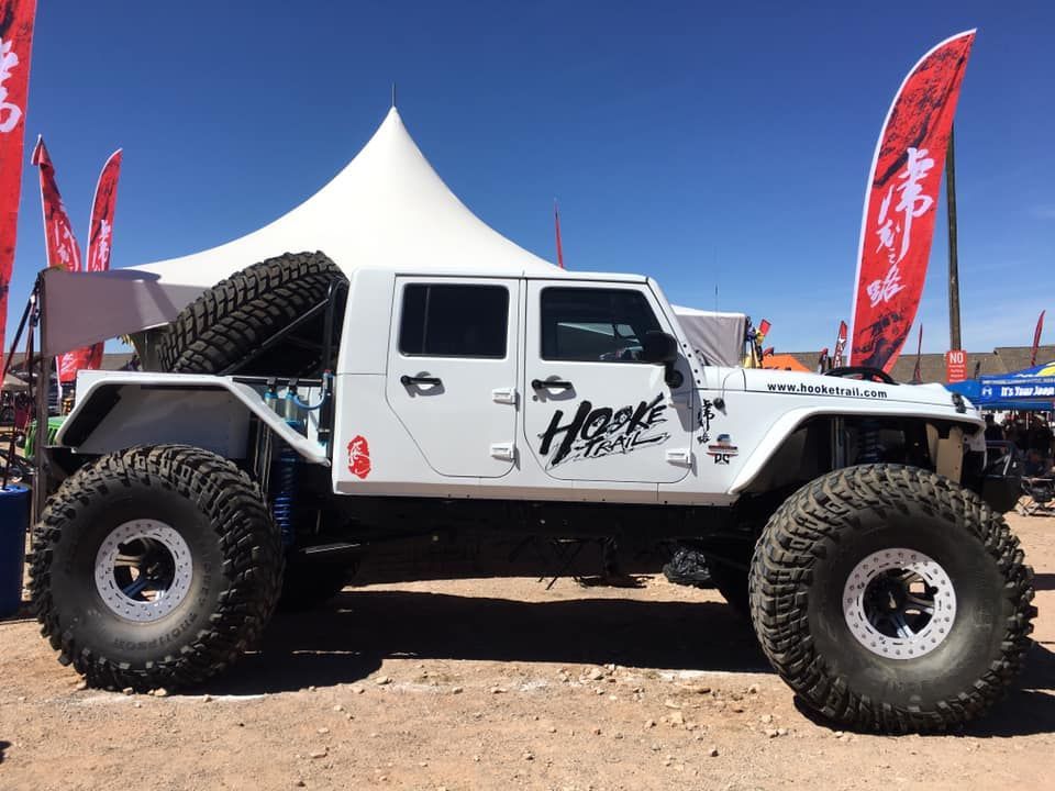 A white modified Jeep Wrangler with massive off-road tires parked at an outdoor event under a blue sky.