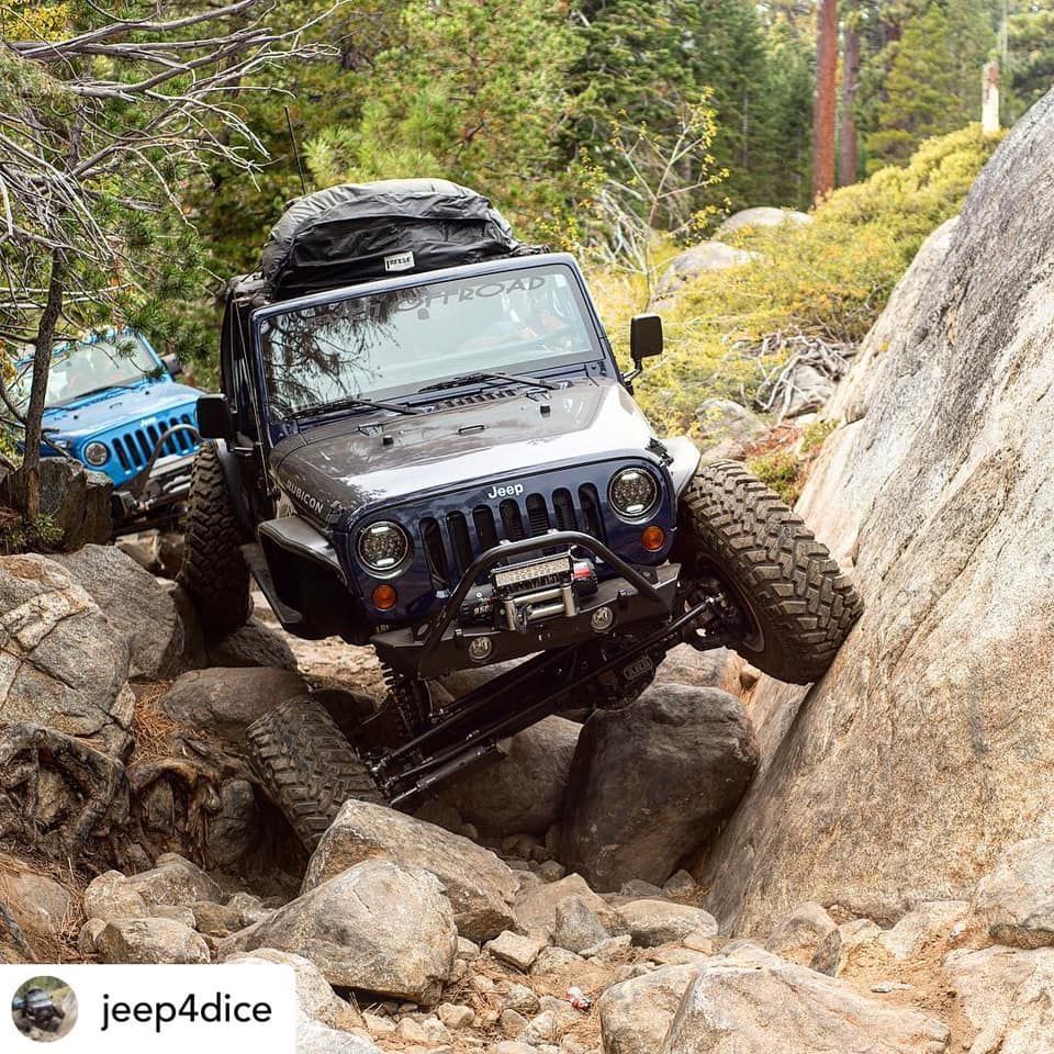 A dark blue Jeep crawls over jagged rocks on a steep trail, with another blue Jeep visible in the background.