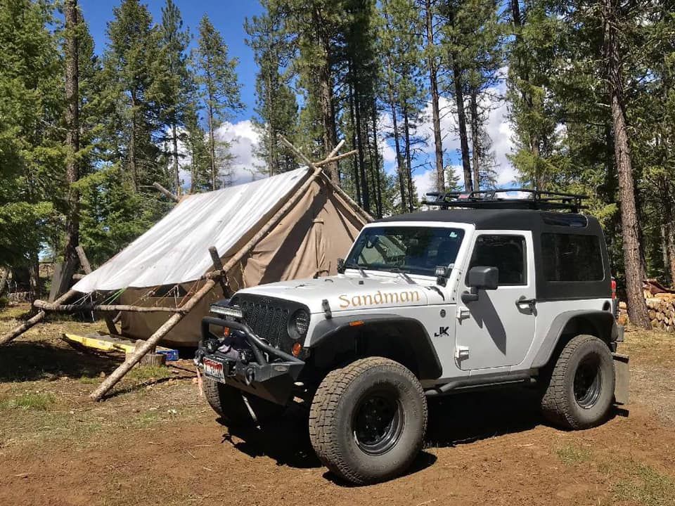 A silver, lifted Jeep parked on a dirt trail in a forest next to a large canvas wall tent.