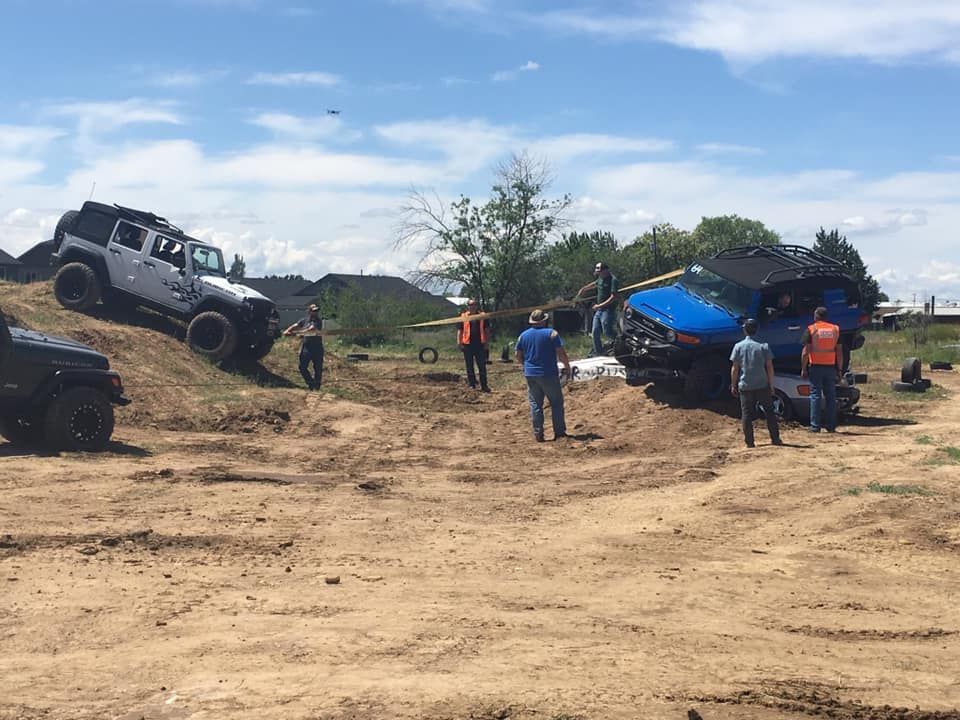A white and a blue off-road vehicle navigate an outdoor dirt course while people in safety vests observe nearby.