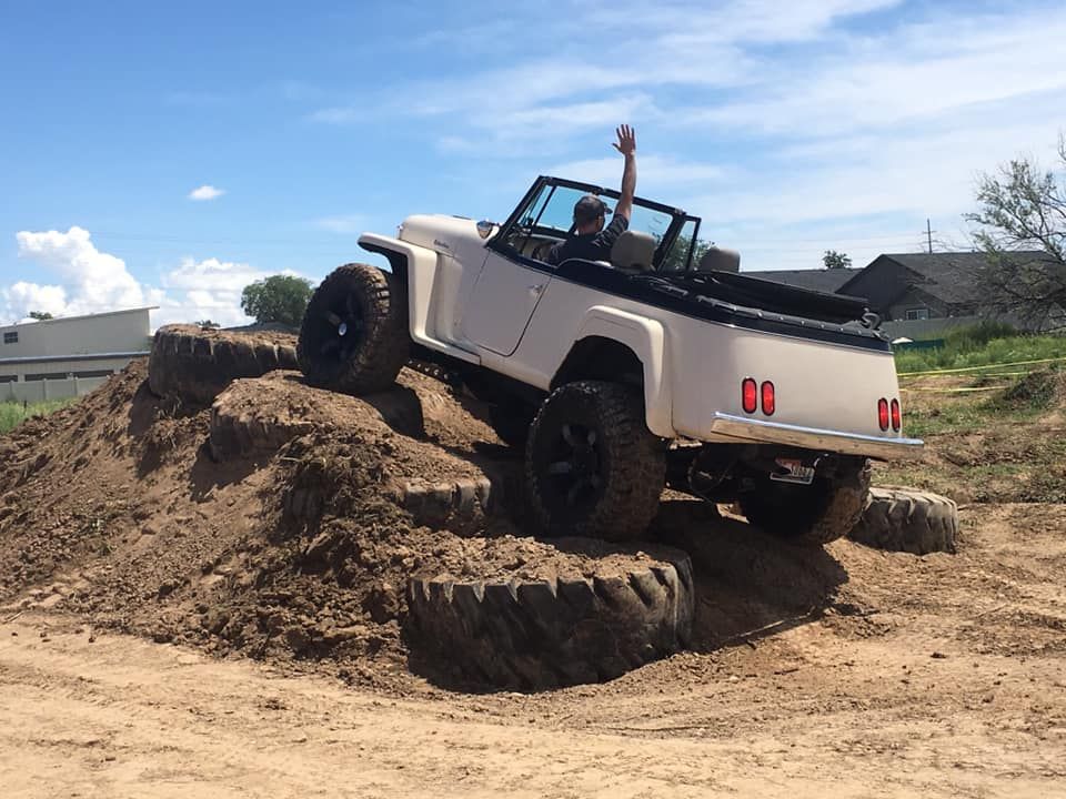 A light-colored off-road vehicle climbs a dirt mound with large tires, while a person inside waves with one hand raised.